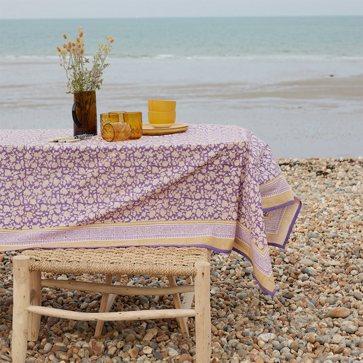 Lifestylebild am Strand: im Vordergrund ein gedeckter Tisch mit Tischdecke Jaya Violine in violett, senfgelb und apricot; im Hintergrund das Meer